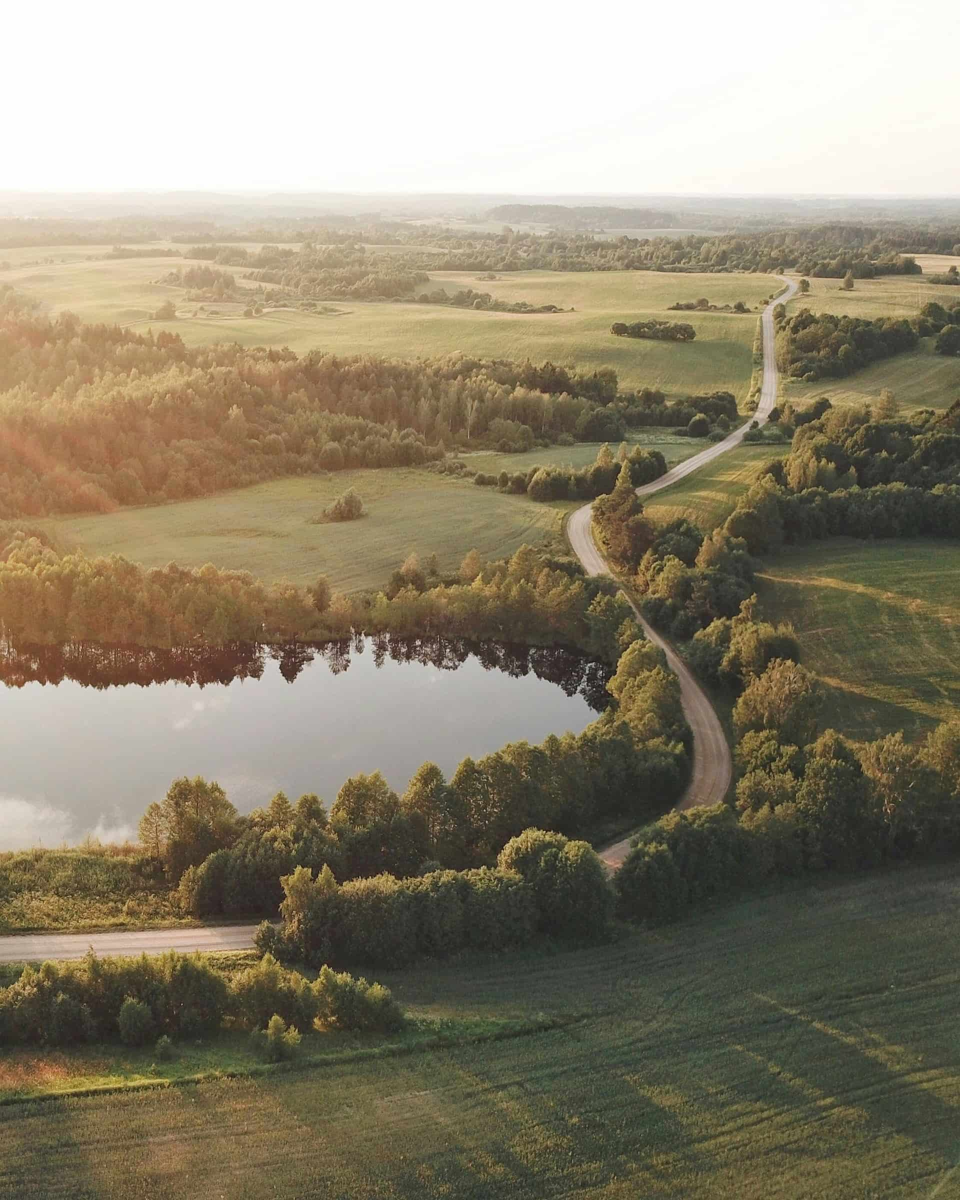 Letecký pohled na harmonicky uspořádanou krajinu s meandrující cestou a rybníkem, obklopený zelenými poli a lesy, ilustruje, jak komplexní pozemkové úpravy přispívají k lepší přístupnosti pozemků, ochraně přírodních zdrojů a zlepšení ekologické stability krajiny.