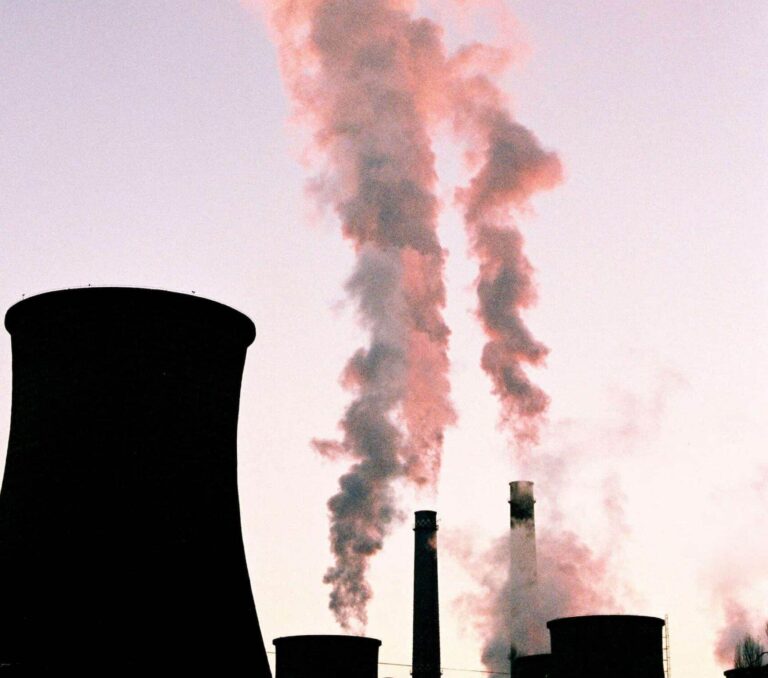 Kouř z komínů elektrárny stoupající do růžově zbarvené oblohy při západu slunce, ilustrující téma uhlíkové stopy a emisí do ovzduší.