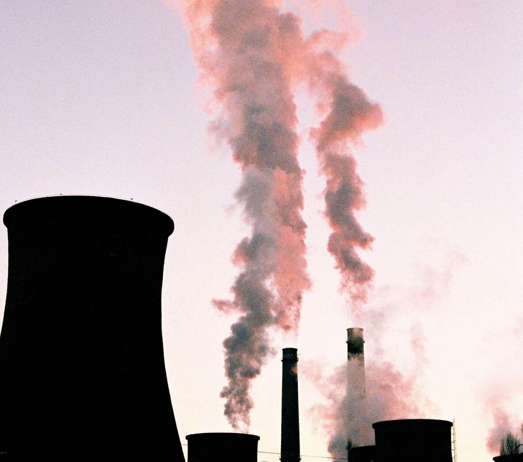 Kouř z komínů elektrárny stoupající do růžově zbarvené oblohy při západu slunce, ilustrující téma uhlíkové stopy a emisí do ovzduší.